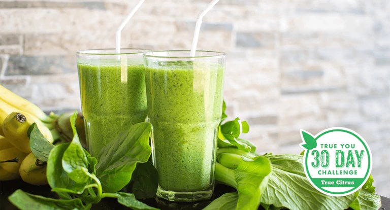 Two glasses of True Lime vanilla protein smoothie next to some leafy greens and bananas.