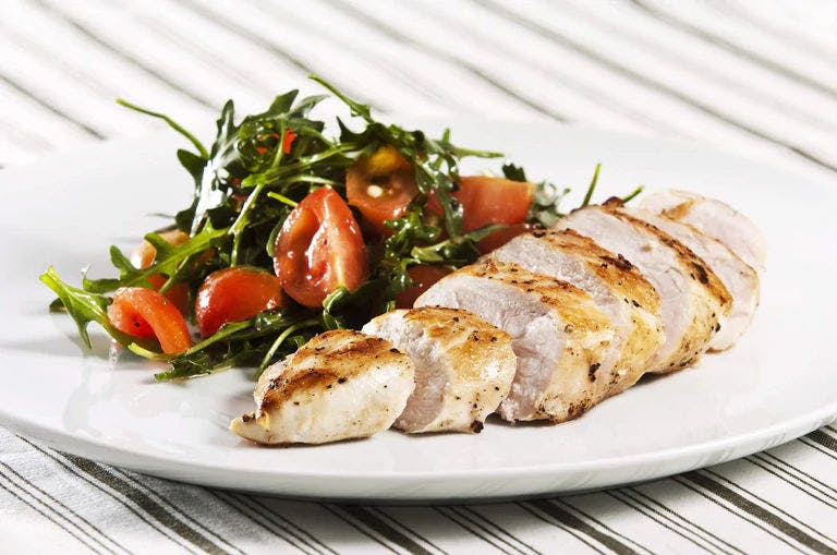 Sliced grilled chicken on plate beside cherry tomato and greens salad