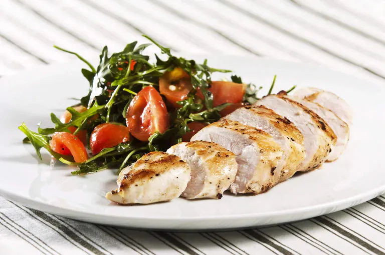 Sliced grilled chicken on plate beside cherry tomato and greens salad