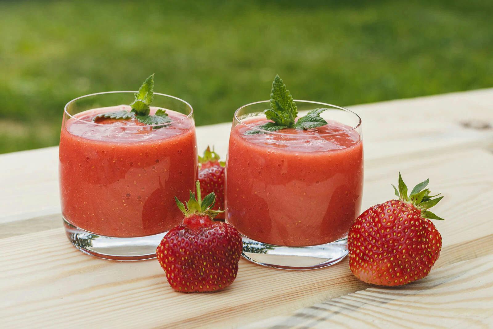 Two glasses of red smoothie on table with strawberries scattered around glasses