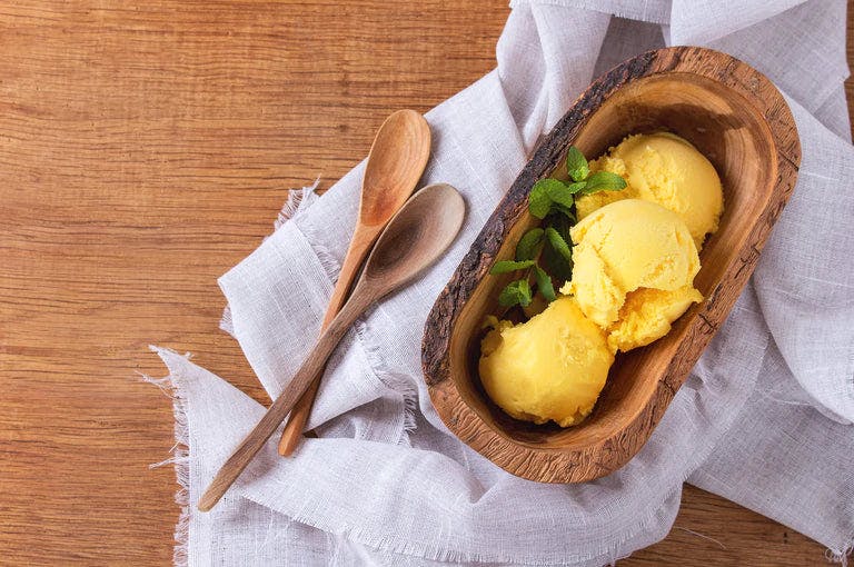 Three scoops of orange sorbet in a wooden bowl with sprigs of mints and wooden spoons to the side