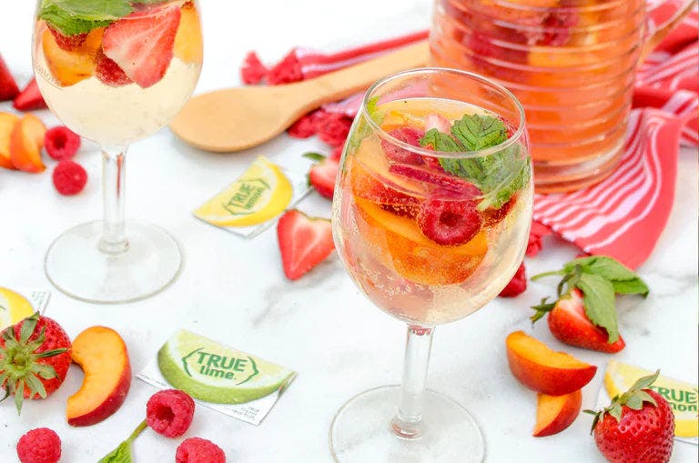 Fruity sangria in wine glasses on table with True Lemon and True Lime packets and various fruit