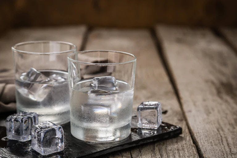 Two glasses of clear liquid with ice cubes in and around glass