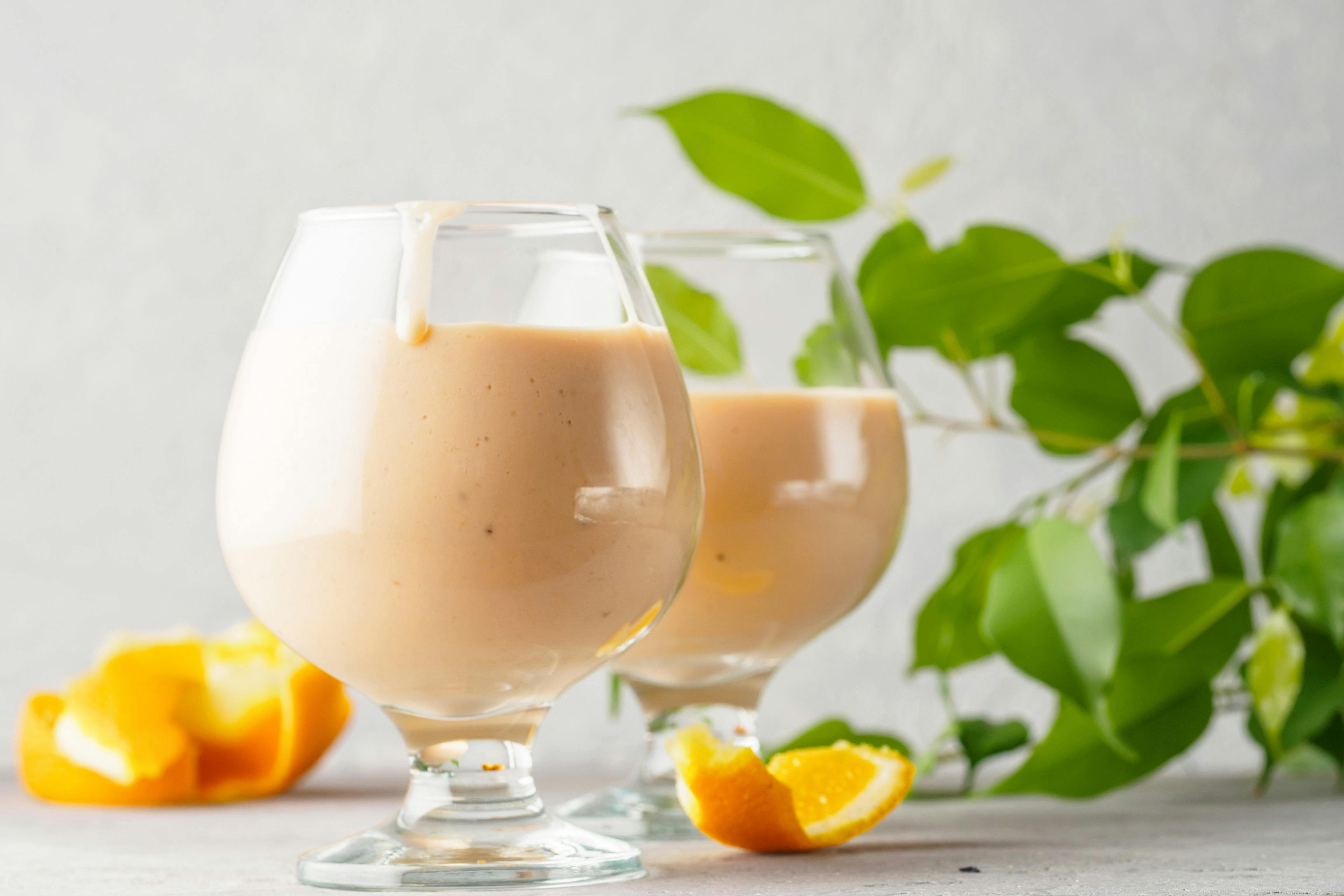 A tasty, creamy Orange Julius in a glass goblet on a countertop with leaves and slices of orange surrounding it.