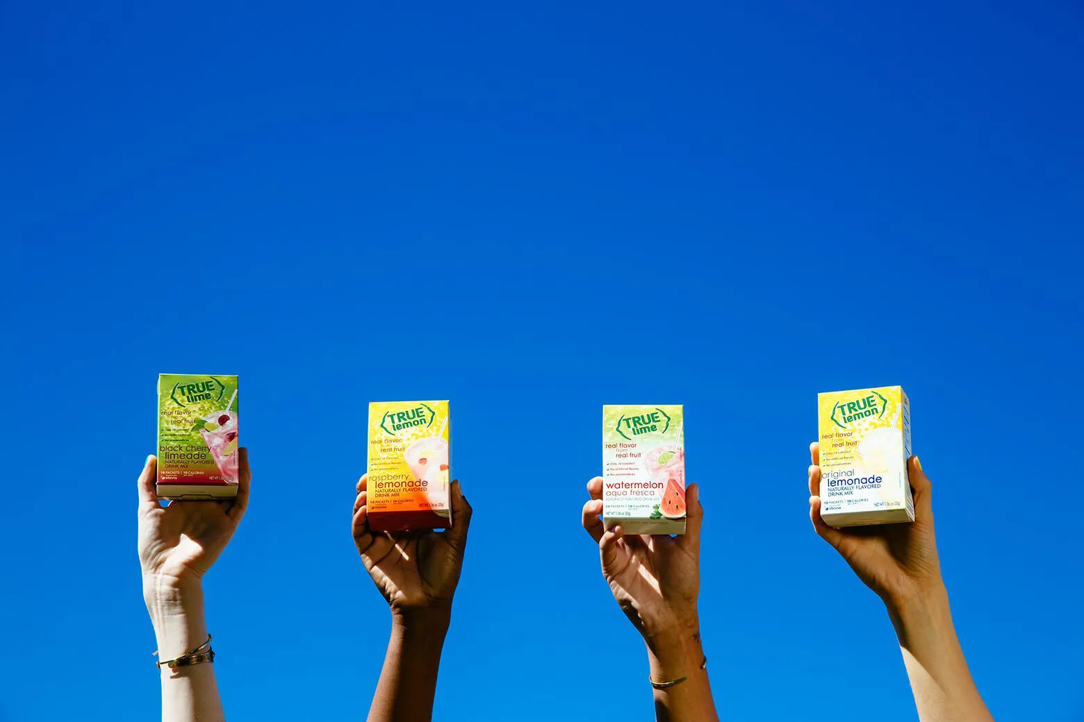 4 hands holding up different True Citrus products against a blue sky.
