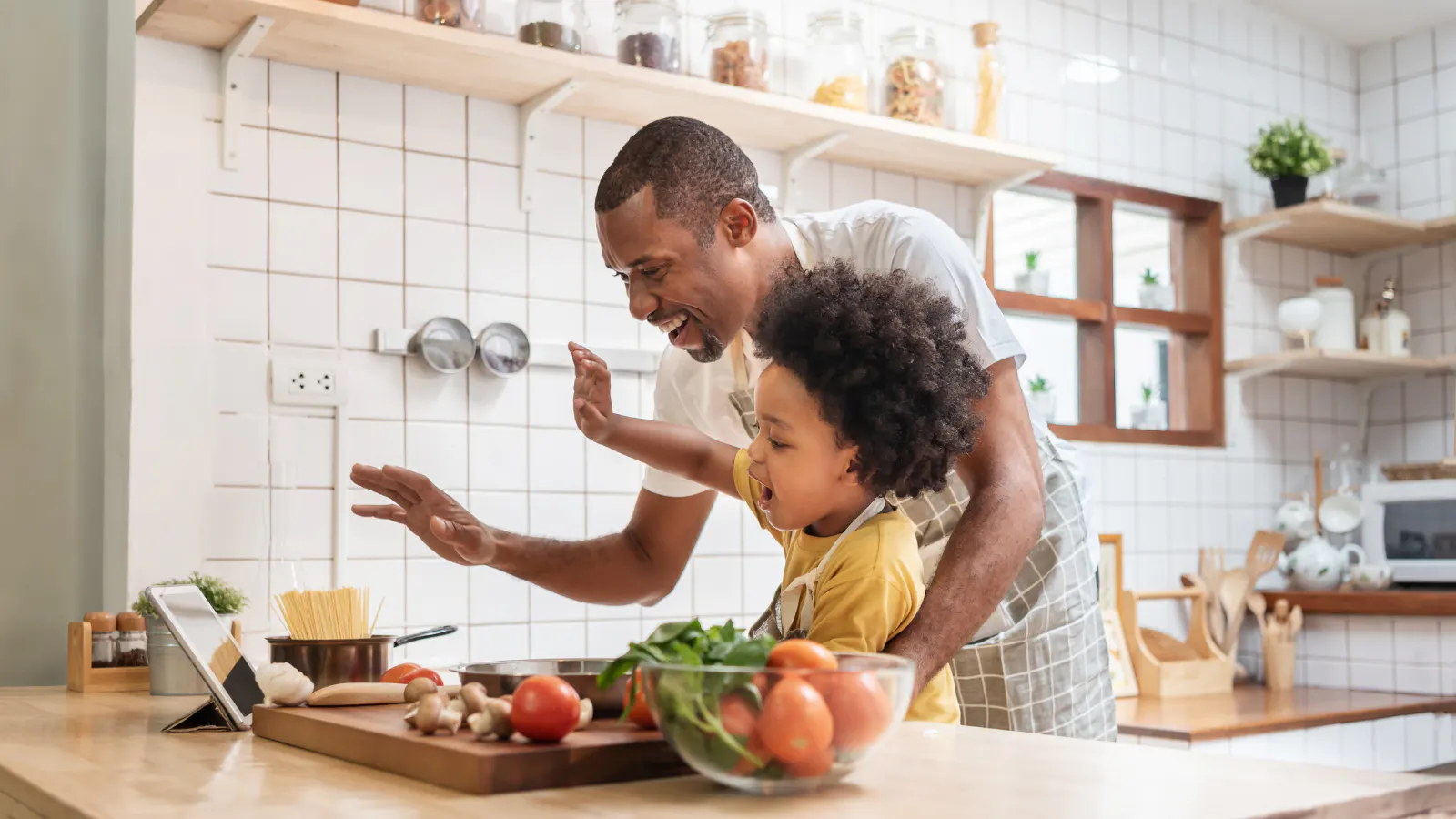 A father an son in the kitchen cooking a meal together. There are vegetables on the counter and they are both laughing.