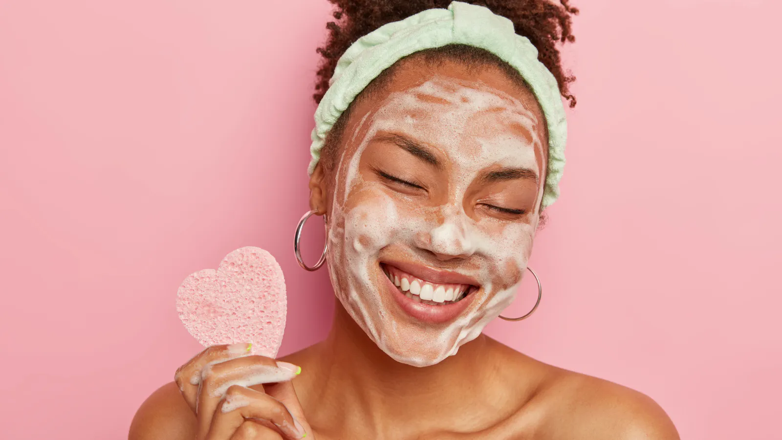 A woman in front of a pink background has face mask on.