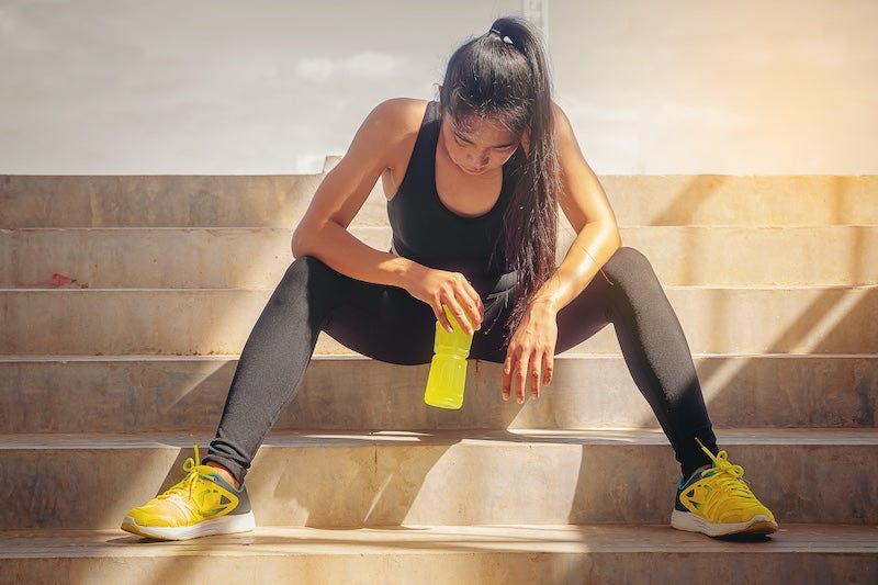 Woman in black athletic clothes sitting down to drink True Lemon after a workout outside