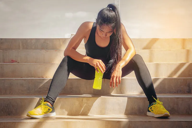 Woman in black athletic clothes sitting down to drink True Lemon after a workout outside