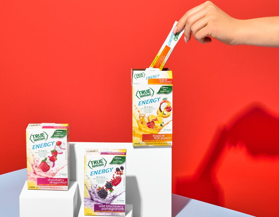 True Citrus Energys on red background with a hand grabbing a packet out of the tropical fruit punch box.