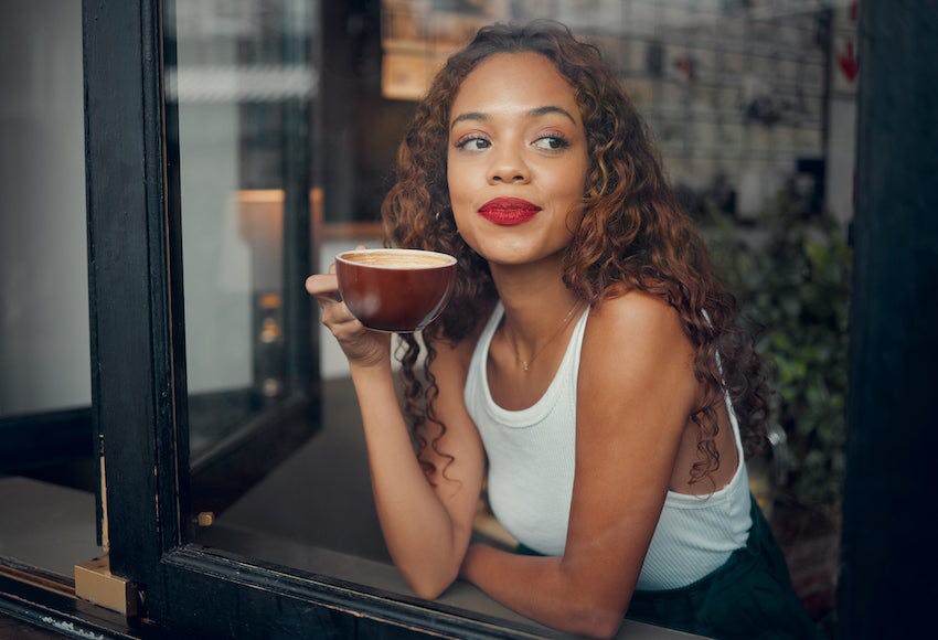 Woman with red lips looking out of the window from a coffee shop holding a maroon mug.